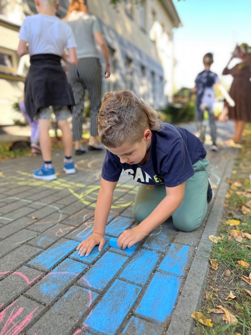 Powitanie Jesieni | Chłopiec na kolanach rysuje niebieską kredą na chodniku, w tle inne dzieci.jpg