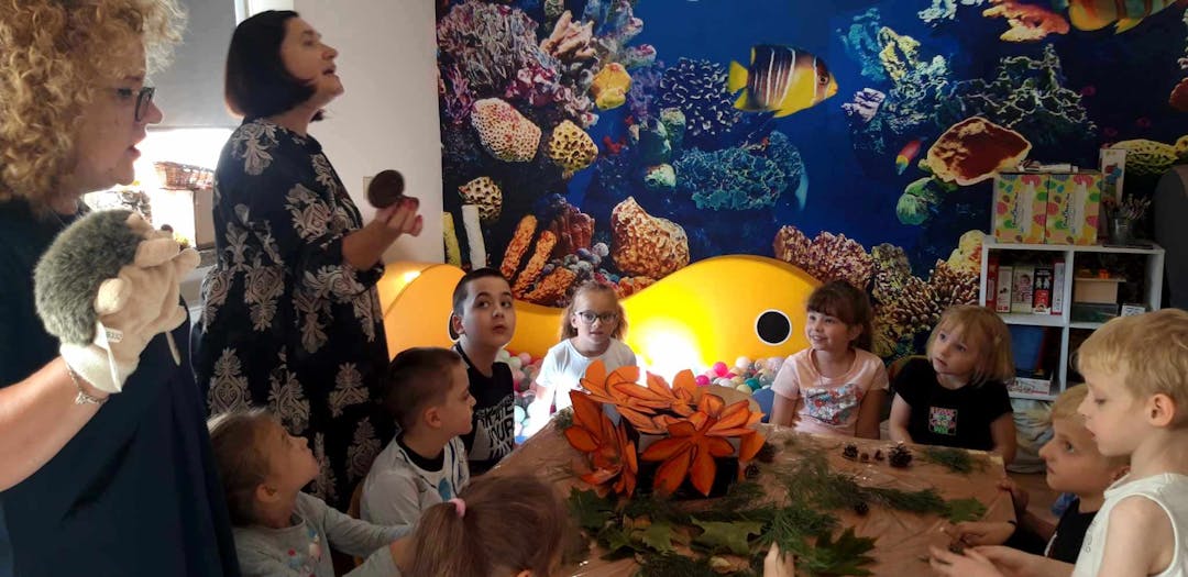 Integracyjne zajęcia w "Krainie uśmiechu" | Dzieci siedza przy stoliku pełnym darow jesieni. Po lewej stronie dwoje nauczycieli z pacynka.jpeg