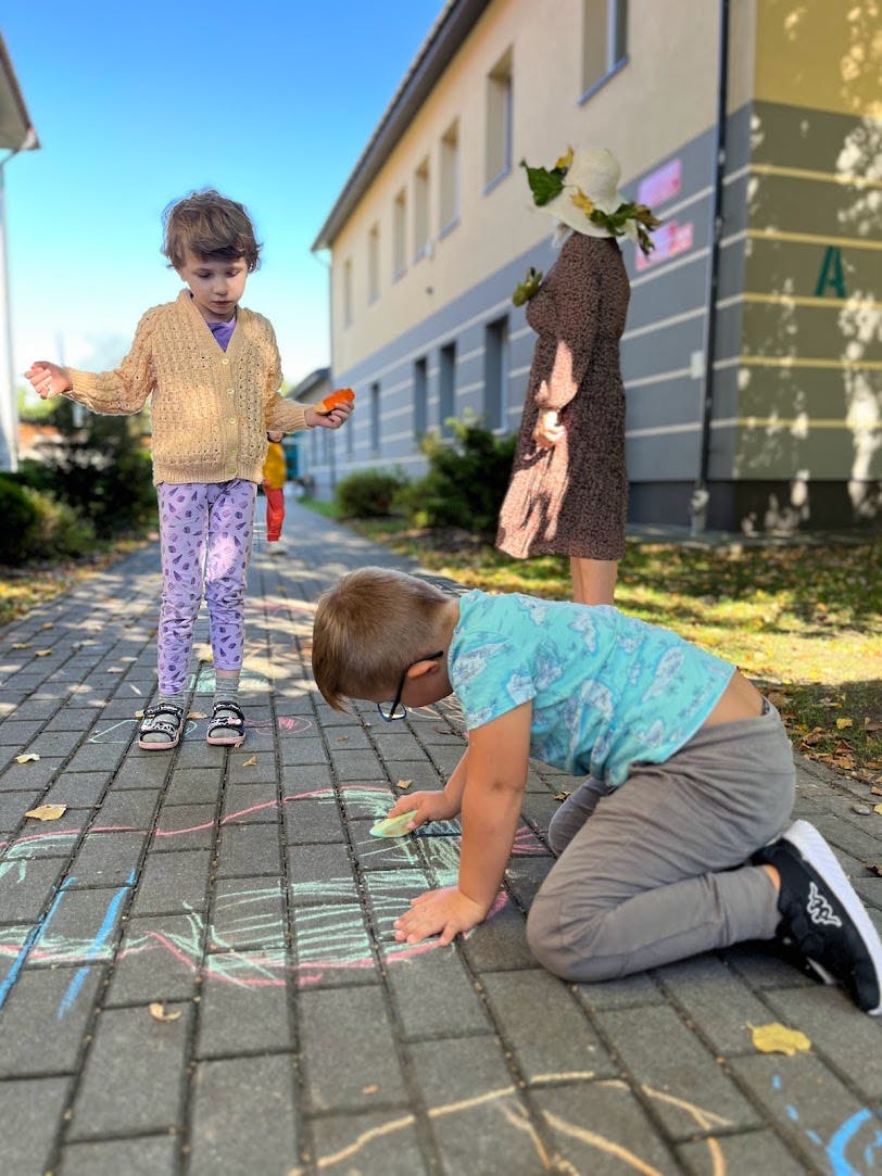Powitanie Jesieni | Chłopiec maluje kredą na chodniku, na drugim planie stoi dziewczynka.jpg