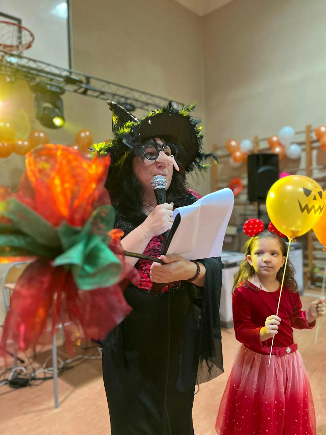 Andrzejki w internacie | nauczycielka przebrana za wróżkę z mikrofonem, obok dziewczynka z balonikiem.jpg