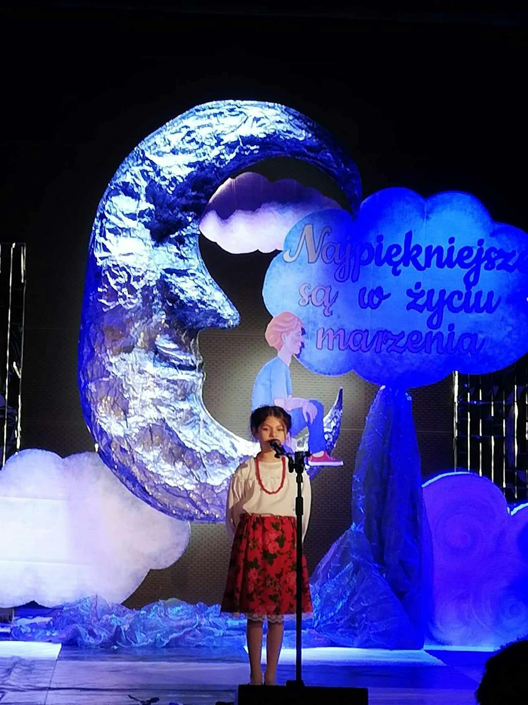 Konkurs recytatorski w Leżajsku | Dziewczynka na czenie przy mikrofonie, w tle dekoracja z księżycem i chmurami.jpg