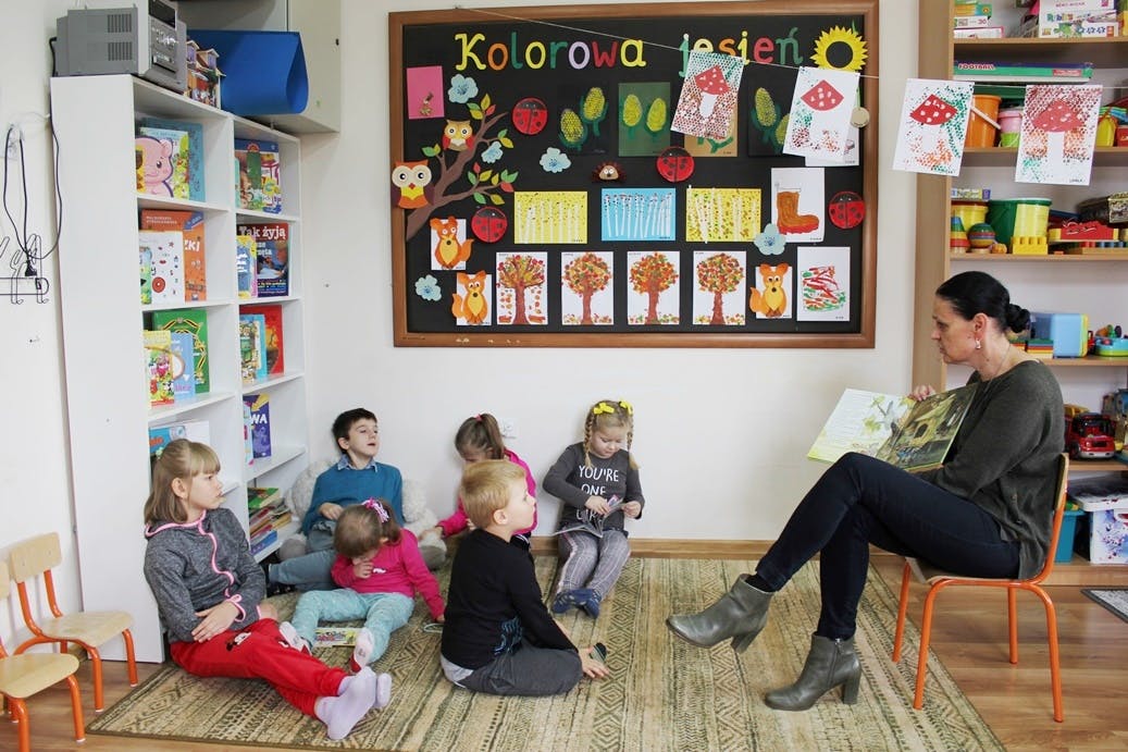 Zrób sobie przerwę na czytanie | Pani siedzi na krzesełku i pokazuje dzieciom obrazek z czytanej bajki. Dzieci siedzą na dywanie naprzeciw pani, słuchają i oglądają_.JPG