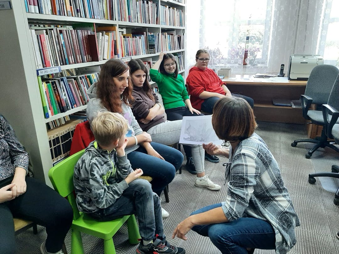 Pozalekcyjne zajęcia w bibliotece | Uczniowie siedzą na krzesłach, nauczyciel pokazuje im obrazek.jpg