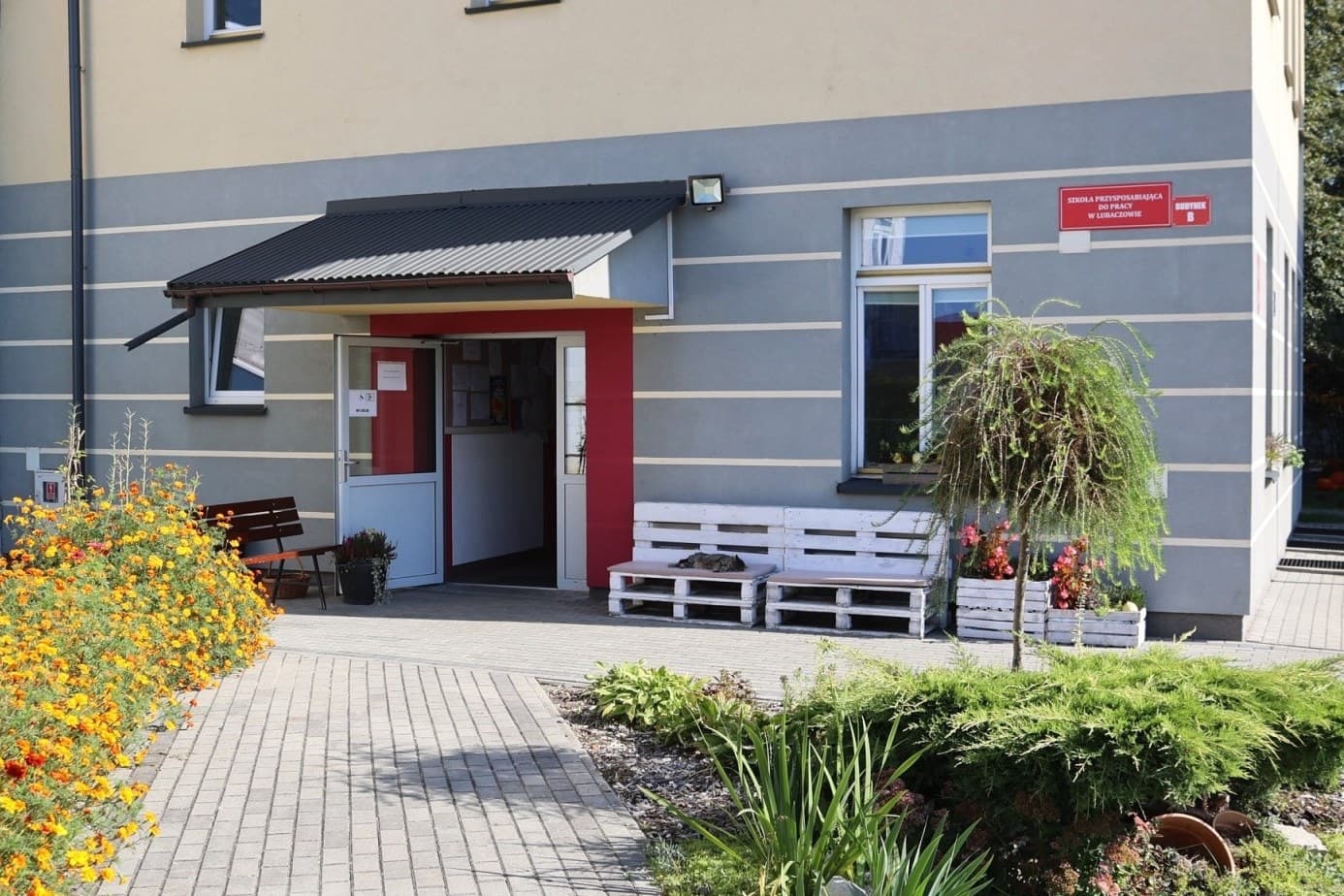 „Przyjazna przystań” | Budynek szkoły, drzwi podkreślone różowymi pasami, obok ławki i kwiaty.jpg