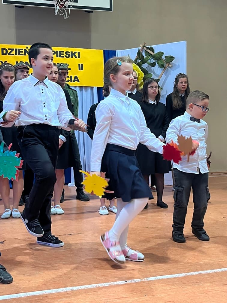 DZIEŃ PAPIESKI | Uczniowie tańczą na scenie z listkami w rękach.jpeg