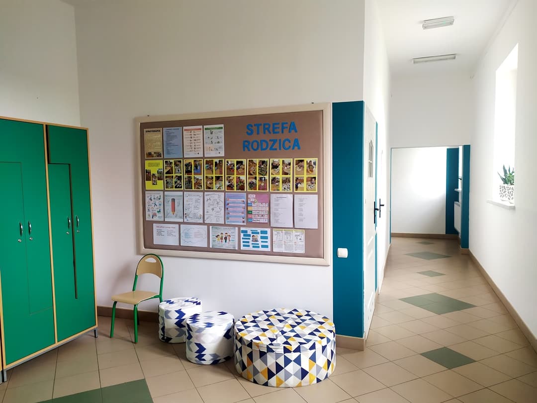 „Przyjazna przystań” | Szatnia i korytasz w szkole, po lewej zielone szafy, na środku pufy i tablica informacyjna.jpg